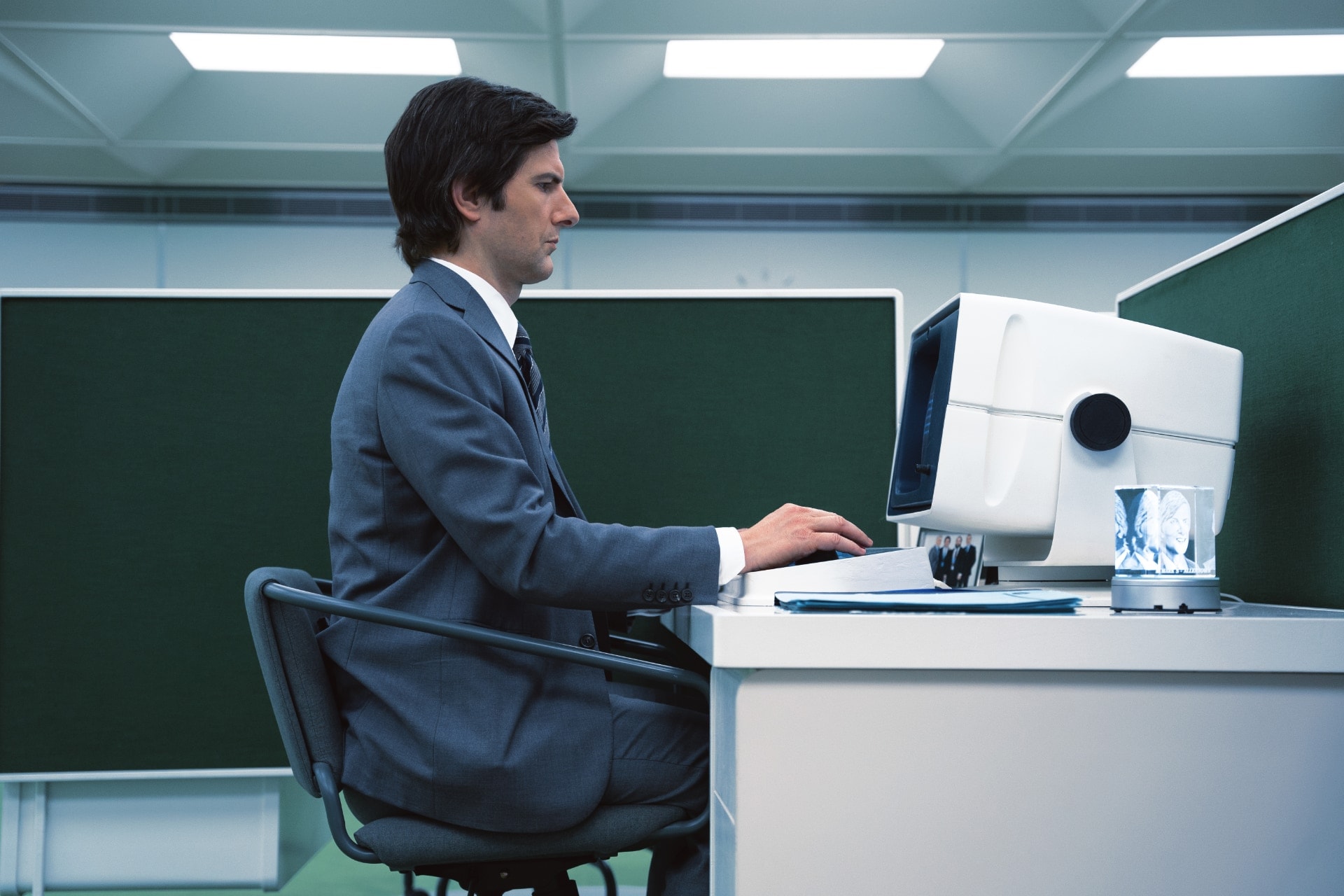 A white man, short hair, sit on his desk, in front an old-fashioned computer, in an old-fashioned office environment.